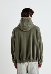 Olajzöld, oversized kapucnis pulóver, húzózsinórral, bordázott mandzsettákkal és szegéllyel. Sima textúrájú, laza szabású, könnyű farmernadrággal viselve.