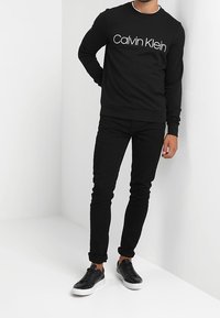 Černá bavlněná mikina s bílým logem "Calvin Klein", doplněná o černé džíny s úzkým střihem a černé tenisky s bílou podrážkou.