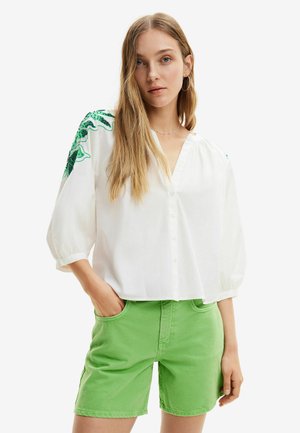 Camicia bianca con bottoni e maniche a sbuffo, con ricami floreali verdi sulle spalle. Abbinata a pantaloni corti in denim verde.