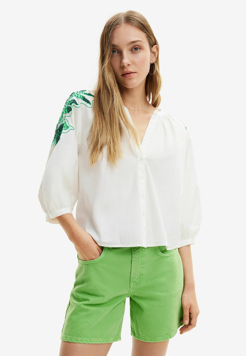 Camicia bianca con bottoni e maniche a sbuffo, con ricami floreali verdi sulle spalle. Abbinata a pantaloni corti in denim verde.