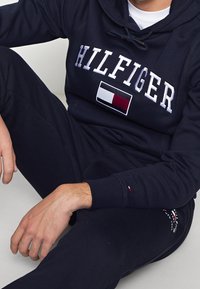Sudadera azul marino con "HILFIGER" bordado en blanco, con un parche de logotipo en color bloque. Mezcla de algodón suave, bolsillo canguro y capucha con cordón.