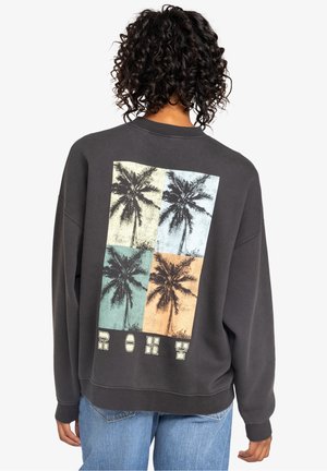 Person mit lockigem Haar, die einen dunklen Sweatshirt mit mehrfarbiger Palmbaum-Grafik und dem Schriftzug „ROXY" auf dem Rücken trägt, kombiniert mit blauen Jeans.