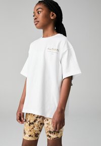 Valkoinen puuvillainen t-paita lyhyillä hihoilla, jossa on kullanvärinen "ALLSAINTS" -logo. Yhdistetty keltaisiin ja ruskeisiin leopardikuosisin shortseihin.