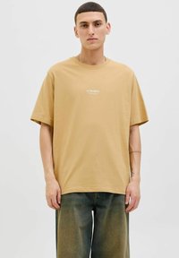 T-shirt en coton beige à coupe relaxe, à manches courtes et avec une marque minimale en texte blanc sur la poitrine; associé à un pantalon ample noir.