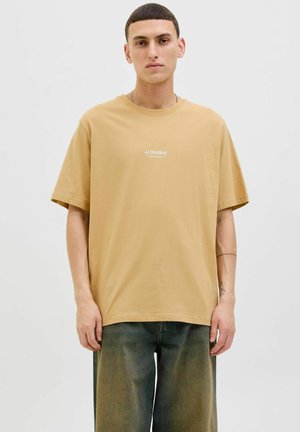 T-shirt en coton beige à coupe relaxe, à manches courtes et avec une marque minimale en texte blanc sur la poitrine; associé à un pantalon ample noir.