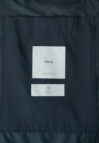 Metka odzieżowa wewnątrz ciemnej tkaniny pokazuje markę „MNG”, rozmiar EU-USA M, wyprodukowana w Wietnamie, z symbolem wodoodporności i tekstem poniżej.