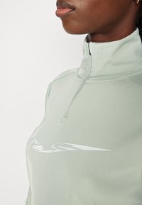 Sudadera de color verde claro con media cremallera que presenta una textura suave y un acento de logo blanco en el pecho. La tela parece elástica y ajustada al cuerpo.