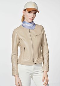 Beżowa skórzana kurtka o slim fit, z zamykanymi kieszeniami i okrągłym dekoltem. Noszona z niebiesko-paskowanym szalikiem i jasnym czapeczką.