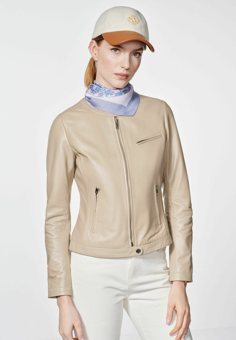 Beżowa skórzana kurtka o slim fit, z zamykanymi kieszeniami i okrągłym dekoltem. Noszona z niebiesko-paskowanym szalikiem i jasnym czapeczką.