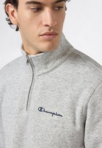 Giovane uomo in felpa grigia chiara con zip a quarter e logo Champion blu ricamato sul petto, che guarda a sinistra.