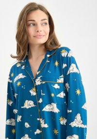 Modrý pyžamový top z měkkého materiálu s potiskem králíčků, mraků a hvězd. Obsahuje kapsu na hrudi a kontrastní žlutou lemovku.
