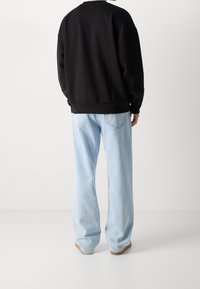 Persona che indossa una felpa nera a maniche lunghe e jeans a gamba larga di colore azzurro chiaro, in piedi con la schiena rivolta verso la fotocamera.