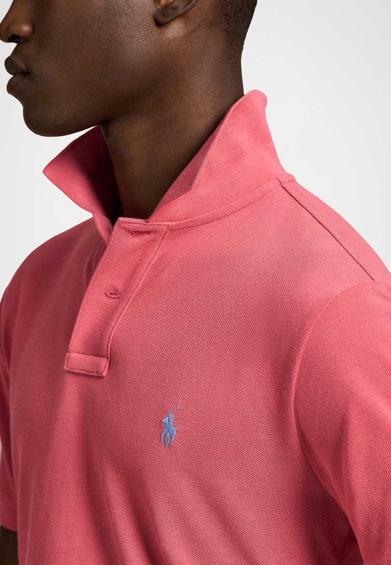 Polo shirt rosa caratterizzato da un tessuto testurizzato, un colletto con bottoni e un logo blu ricamato sul petto, che mette in mostra un design aderente.