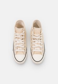 Wysokie trampki wykonane z beżowej, teksturowanej tkaniny, z białymi sznurówkami, gumowym noskiem i czarnym akcentem przy podeszwie.