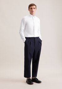 Camicia bianca aderente con colletto, chiusura frontale con bottoni, abbinata a pantaloni a pieghe blu scuro e scarpe nere con lacci.