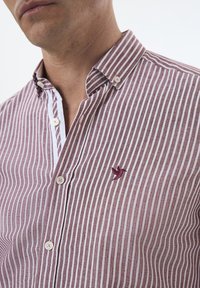 Camisa de hombre de botones con rayas burdeos y blancas, cuello corto y logo de pájaro bordado en el pecho. Confeccionada en tela ligera.