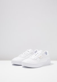 Zapatillas blancas con parte superior de cuero liso, detalles perforados, cuello acolchado y suela de goma texturizada. Incluye un logotipo con acento dorado.