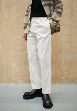 Persona con pantalones blancos de pierna recta, botas negras gruesas, camisa negra y chaqueta a cuadros beige, de pie frente a una pared beige texturizada.