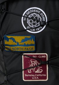 Black fabric backpack featuring three embroidered patches: a round patch with a geodesic dome, a blue and gold patch with mountain graphics, and a maroon patch depicting climbing.
