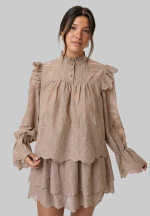 Młoda kobieta ubrana w beżową koronkową sukienkę z falbanami na ramionach, zapiętym na guziki wysokim kołnierzem, rękawami dzwonkowymi oraz warstwową spódnicą, stoi na tle szarego tła.