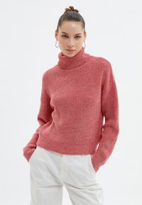 Pull en maille rouge, épais, avec un col roulé haut et des manches longues. Le tissu a une apparence texturée, offrant un look confortable.
