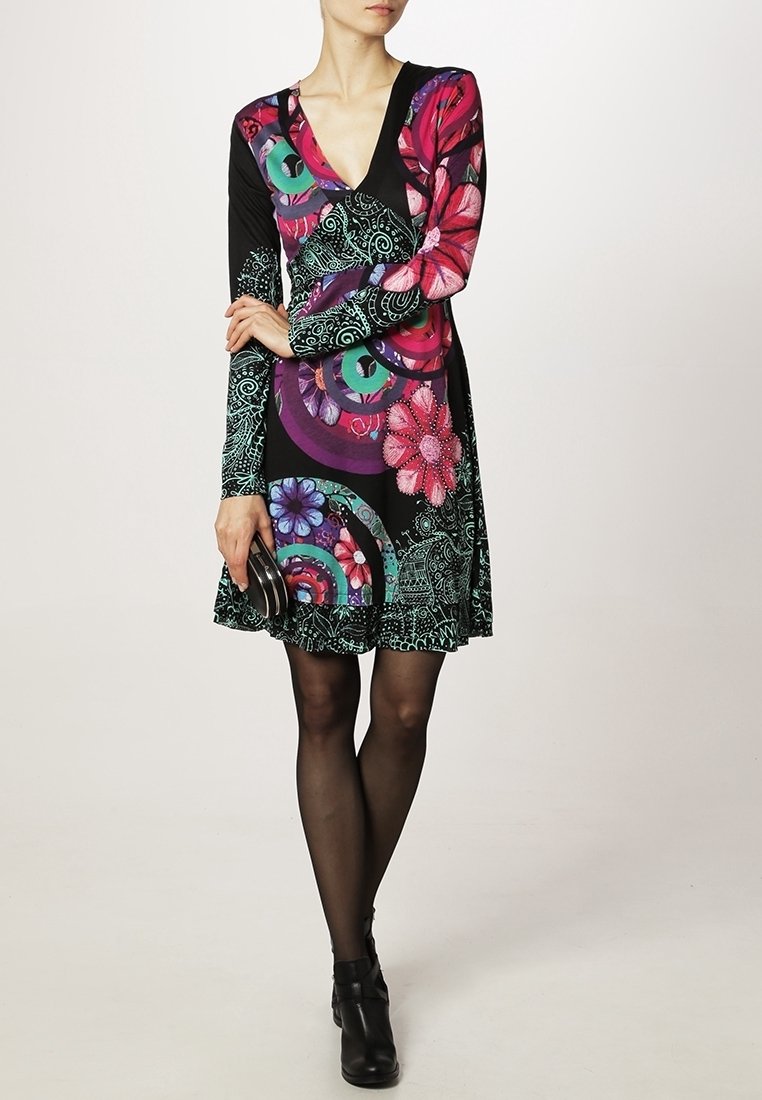 Nainen mustissa nilkkabootseissa, läpinäkyvissä mustissa sukkahousuissa ja pitkähihaisessa mekossa, jossa on värikkäitä ympyrä- ja kukkakuoseja, pitää käsissään pientä mustaa clutch-laukkua.