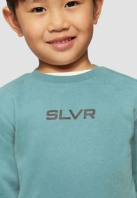 Lichtblauwe sweatshirt met ronde hals. Bevat een donkergrijze "SLVR" logo dat prominent op de borst is afgebeeld. Zachte textuur.