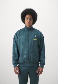 Giacca sportiva color teal con zip frontale, motivo tonale, colletto alto e dettagli del logo in verde chiaro e giallo. Indossata con pantaloni coordinati.