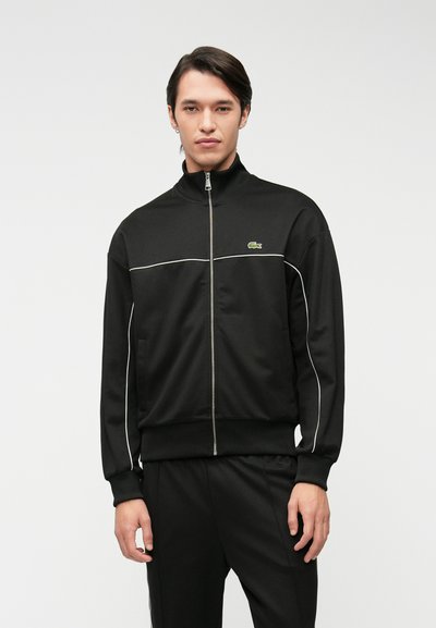 Veste noire zippée en tissu lisse, avec un passepoil blanc sur la poitrine et un petit logo vert sur le côté gauche. Design avec col montant.