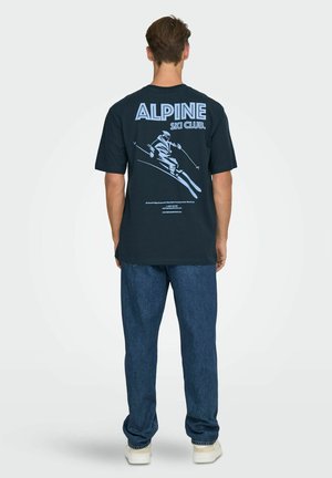 Navy blue cotton t-shirt featuring "ALPINE SKI CLUB" text and a skiing graphic on the back; paired with blue denim pants and white shoes.