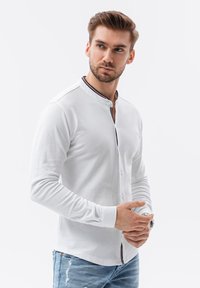 Camisa blanca de manga larga con una tapeta abotonada y detalle de raya azul marino en el cuello, combinada con jeans azules desgastados.