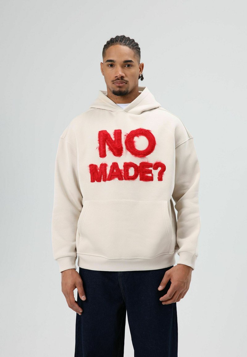 Roomwitte capuchontrui met rode pluizige tekst "NO MADE?" op de voorkant, kangoeroezak en geribbelde manchetten, gecombineerd met donker denim broek.