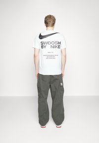 Vit bomullströja med svart text "SWOOSH BY NIKE" och logotyp på baksidan, tillsammans med löst sittande olivgröna cargobyxor.