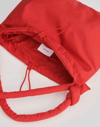 Borsa in tessuto rosso con una texture morbida, chiusura a coulisse, manici imbottiti e una tasca interna con zip. L'etichetta appare all'interno.