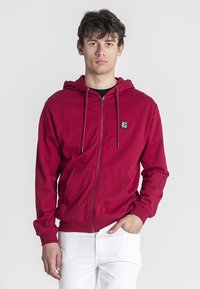 Crvena majica s dugim rukavima zatvarač, izrađena od mekanog materijala, sa prednjim zatvaračem, duplim bočnim džepovima i kapuljačom sa vezicom. Logotipna oznaka na prsima.