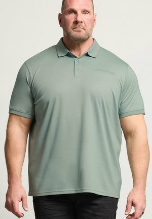 Mann mit kurzen Haaren und Bart, der ein hellgrünes Polohemd mit kurzen Ärmeln und schwarze Hose trägt und mit dem Gesicht nach vorne vor einem einfarbigen Hintergrund steht.