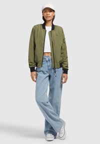 Giacca bomber verde oliva con dettagli neri, materiale leggero, tasche con zip, abbinata a jeans a gamba larga azzurri e sneakers bianche.