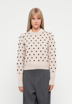Pull en tricot rose clair avec des pois noirs, des bords côtelés et un col rond. Porté avec un pantalon taille haute gris.