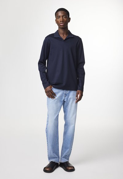 Polo à manches longues bleu marine avec col, associé à un jean bleu clair et des sandales noires. La chemise présente une texture simple et lisse.