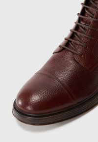 Bruine leren laars met textuur en een ronde neus, voorzien van vetersluiting en tonale veters, aangevuld met een leren zool en decoratieve stiksels.