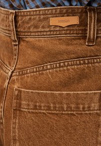 Bolsillo de jeans de mezclilla marrón con trabillas para cinturón y etiqueta de cuero con la marca "sessùn" cosida sobre el bolsillo, combinado con camisa azul con cuadros.