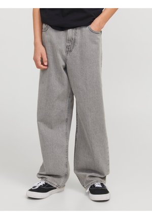 Person iført løse grå jeans og sorte sneakers med hvide snørebånd, den ene hånd i lommen, står på en ensartet lys baggrund.