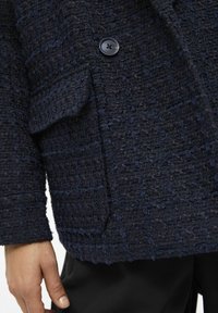 Blazer texturé bleu marine avec un motif tissé, deux poches avant et une fermeture à bouton noir. Le tissu est épais et substantiel.