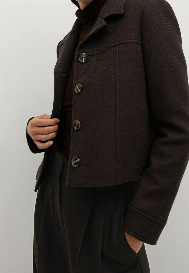 Personne portant une veste courte boutonnée marron foncé sur un pull à col roulé foncé, une main dans la poche et l'autre ajustant la veste.