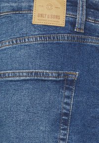 Donkerblauwe spijkerbroek met een gestikte achterzak, voorzien van een tan leren label met "ONLY & SONS" en decoratieve stiksels.