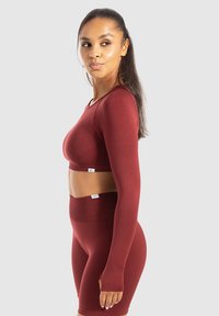 Vrouw met lang haar die een aansluitende maroon lange mouwen crop top en hoge taille fietsshorts draagt, staande opzij tegen een effen achtergrond.