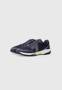 Tênis de atletismo em azul marinho com detalhes em branco e a marca EA7. Possui um cabedal texturizado, fechamento com atacadores e uma sola verde limão com um padrão de aderência.