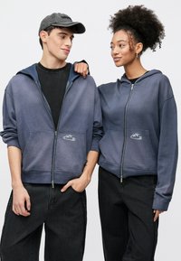 Uniszex, cipzáros kapucnis pulóverek kék színátmenetben, kenguruzsebbel és hímzett mintával. Fekete széles szárú nadrággal párosítva.