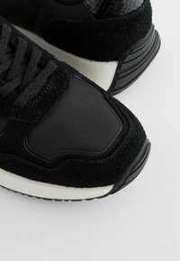 Schwarze Sport-Sneaker, die eine Kombination aus glatten und strukturierten Materialien aufweisen, mit einer gepolsterten Sohle und filigranen Details im Oberdesign.