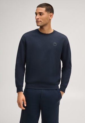 Mann mit kurzen Haaren, der einen marineblauen Pullover und passende Hosen trägt, steht mit einer Hand in der Tasche und schaut nach links vor einem neutralen Hintergrund.